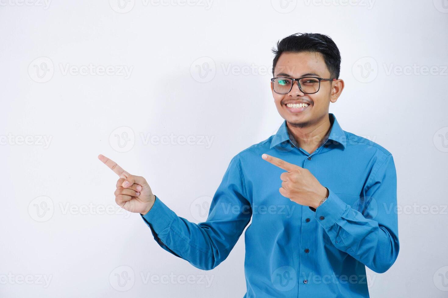 Handsome asian male employee with glasses smiling in a pose finger pointing to the side wearing blue shirt isolated on white background photo