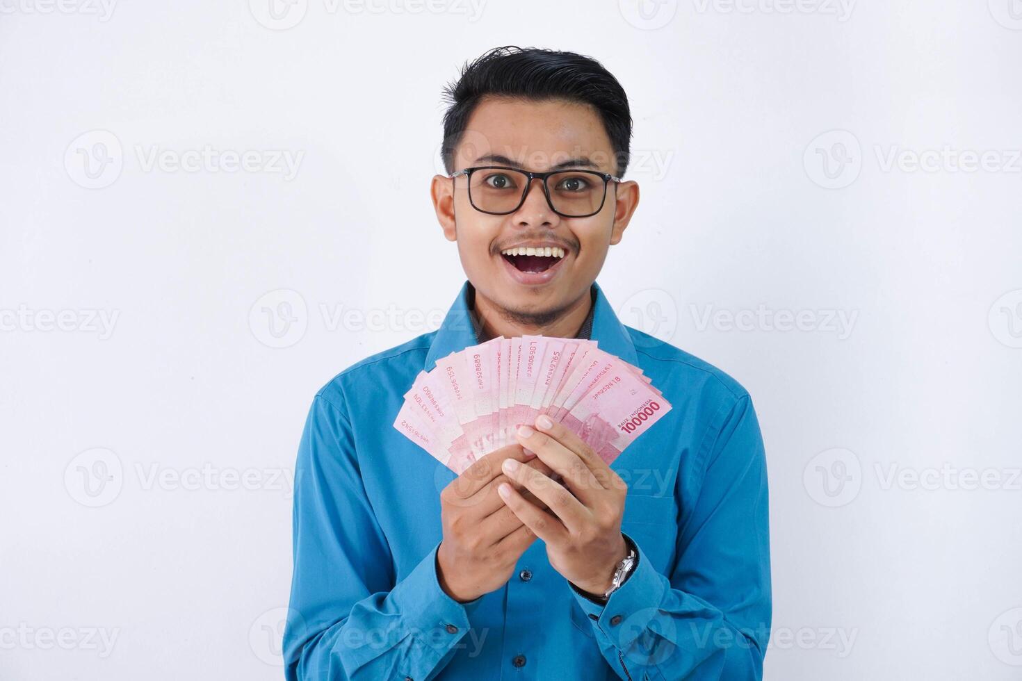 surprised or shocked handsome young asian businessman in wearing blue shirt and glasses holding fan of indonesia banknotes in hands isolated on white background photo