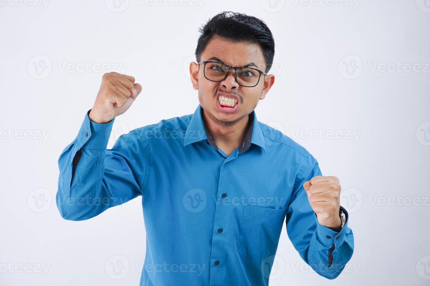 Angry aggressive young asian man with glasses wearing blue shirt with ferocious expression and mad raising fists frustrated isolated on white background photo