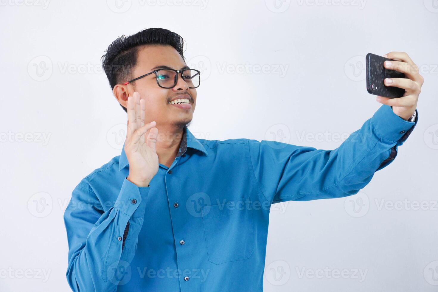 smiling or happy asian man with glasses holding smartphone for selfie photo wearing blue shirt isolated on white background