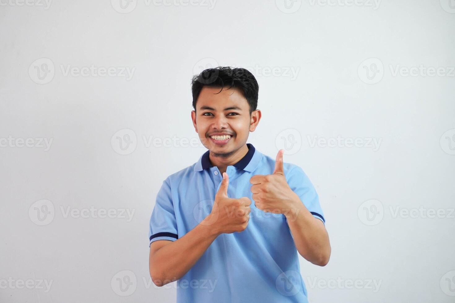 retrato de alegre asiático hombre en vistiendo azul t camisa sonriente y demostración pulgares arriba a cámara aislado terminado blanco antecedentes foto