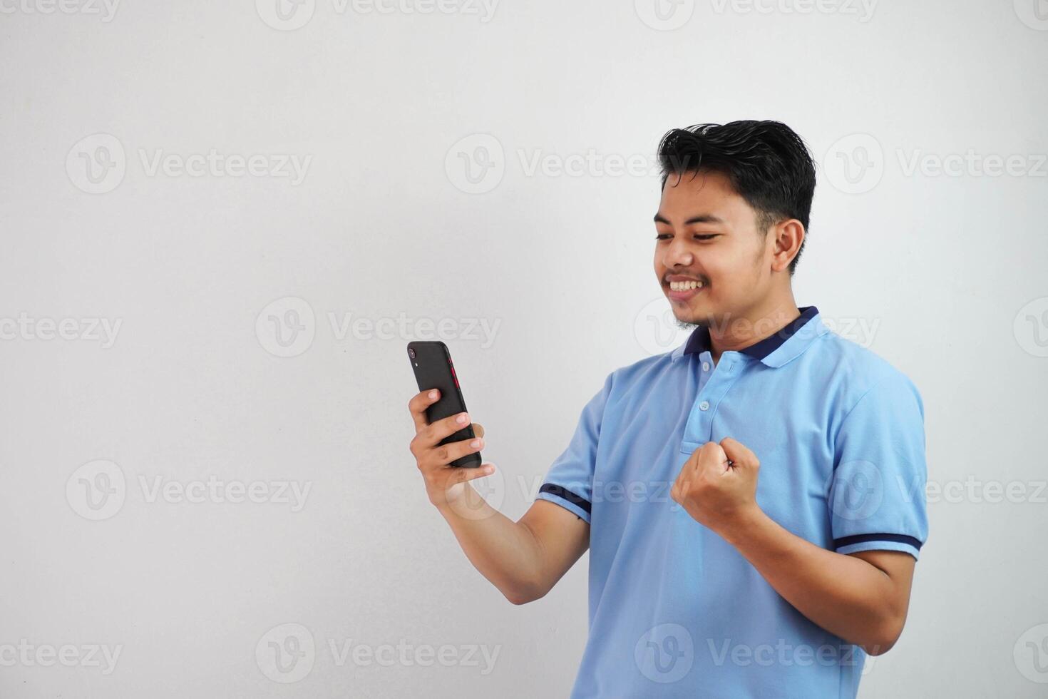 excited or happy portrait young asian man holding phone with clenched fists wearing blue polo t shirt isolated on white background photo