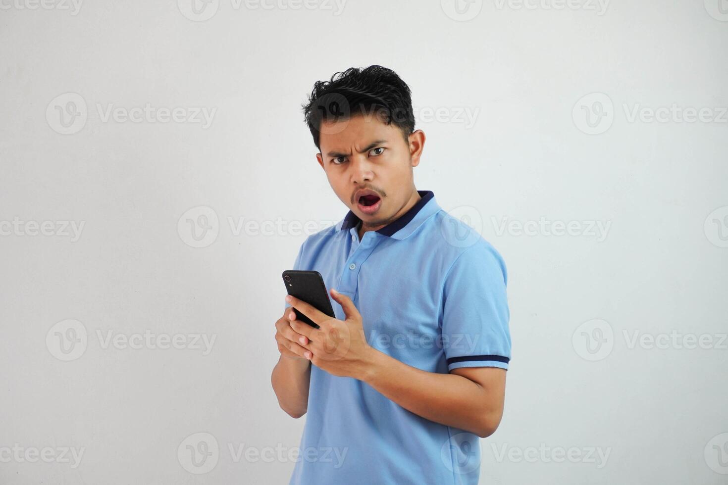 joven asiático hombre con asombrado expresión mirando insatisfecho con irritado cara mientras apertura su boca y participación teléfono vistiendo azul t camisa aislado en blanco antecedentes foto