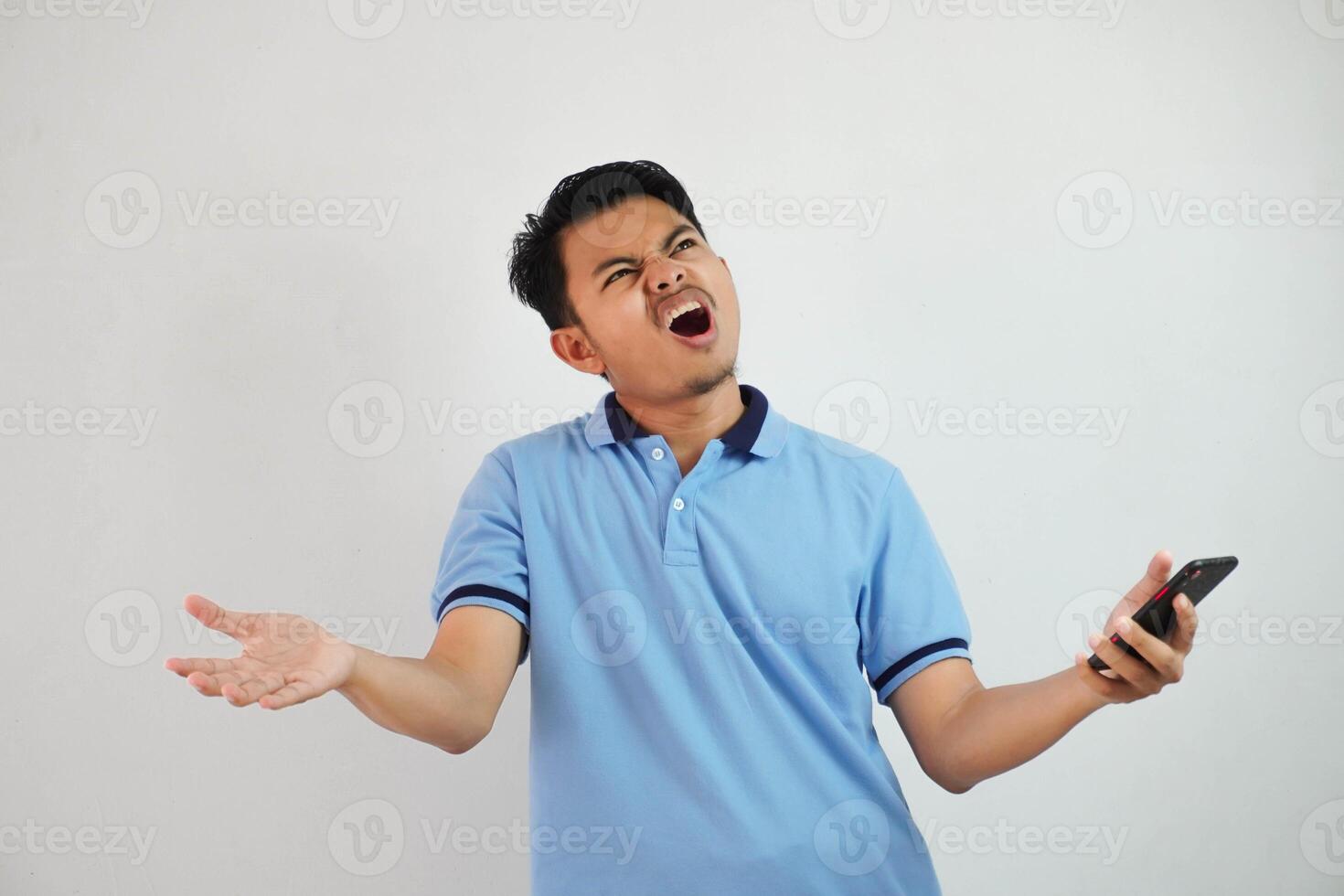 young Asian man with astonished expression looking dissatisfied with annoyed face while opening his mouth and holding phone wearing blue t shirt isolated on white background photo