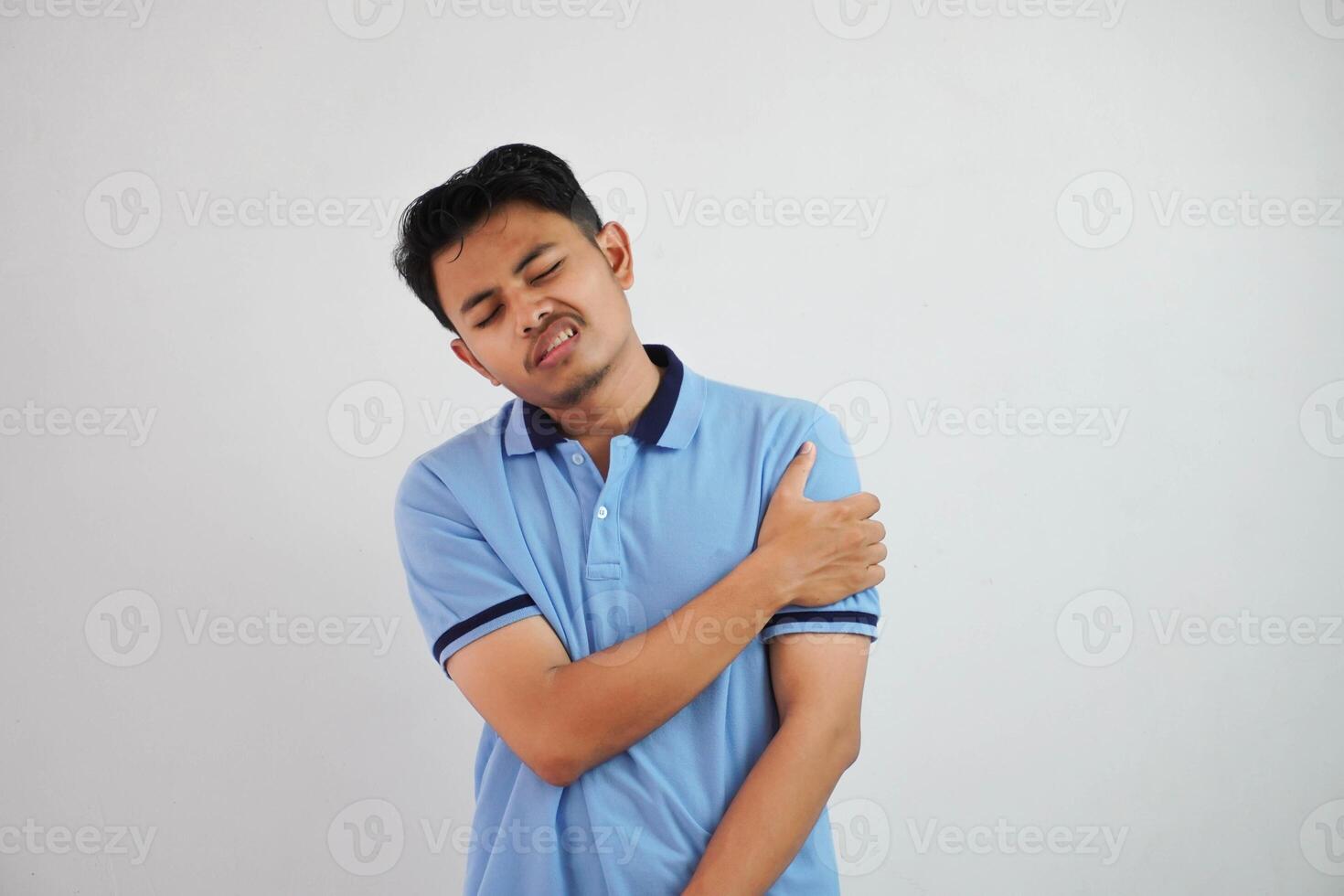 joven asiático hombre azul t camisa gesto dolor su hombro o brazo en dolor aislado en blanco antecedentes foto