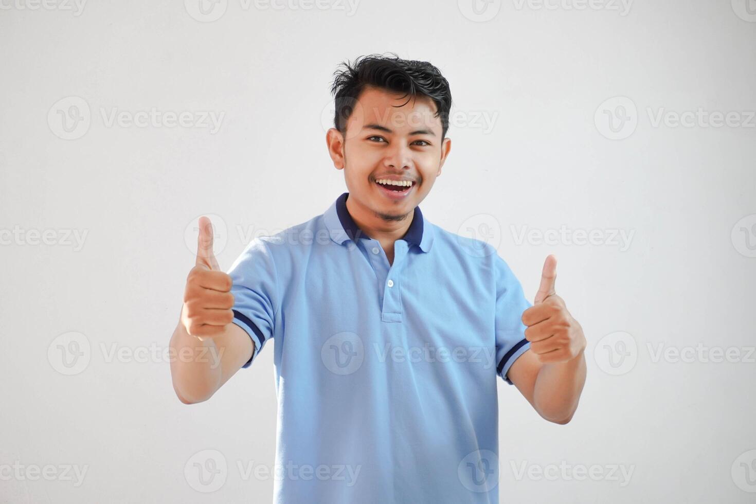 Portrait of cheerful asian man in wearing blue t shirt smiling and showing thumbs up at camera isolated over white background photo