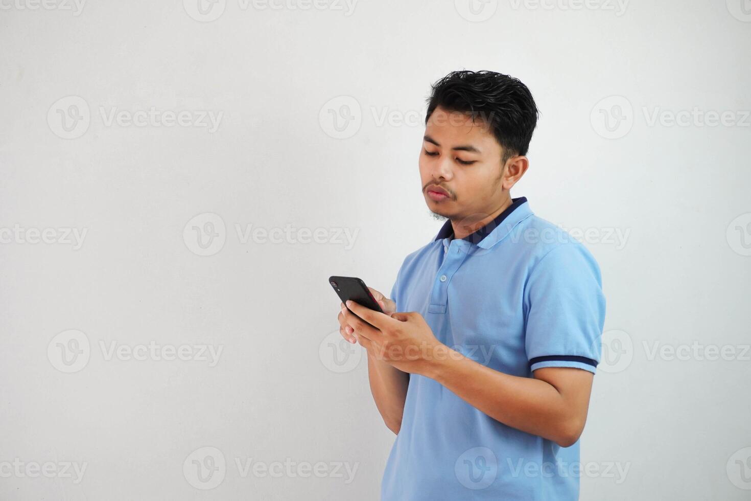 joven asiático hombre en pie participación un teléfono con un enojado y desaprobando expresión vistiendo azul t camisa aislado en blanco antecedentes foto