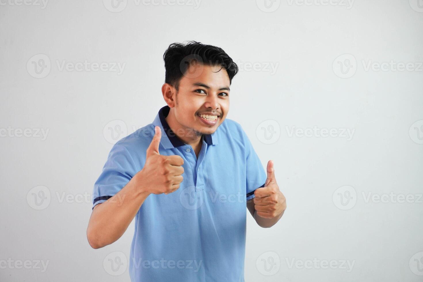 Portrait of cheerful asian man in wearing blue t shirt smiling and showing thumbs up at camera isolated over white background photo