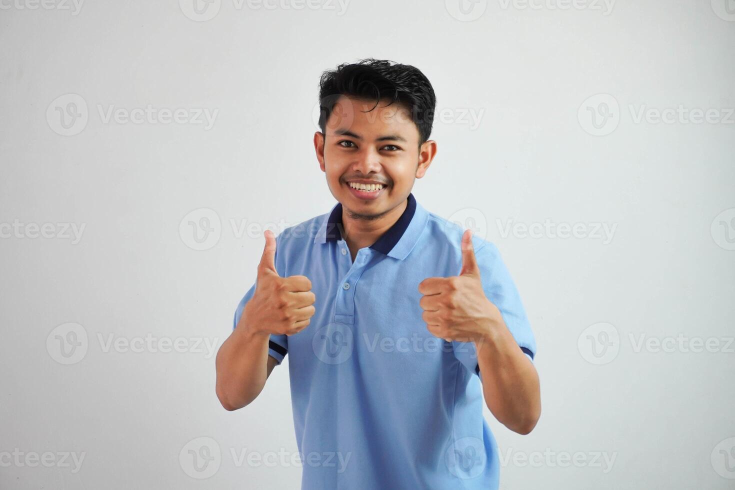 retrato de alegre asiático hombre en vistiendo azul t camisa sonriente y demostración pulgares arriba a cámara aislado terminado blanco antecedentes foto