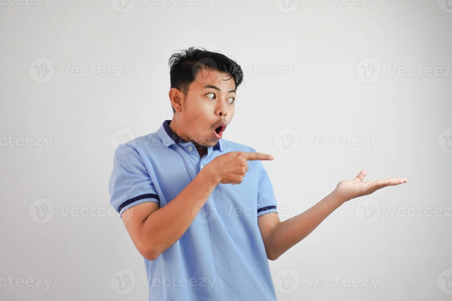 conmocionado asiático hombre con un abierto mano con dedos señalando a el lado vistiendo azul t camisa aislado en blanco antecedentes foto