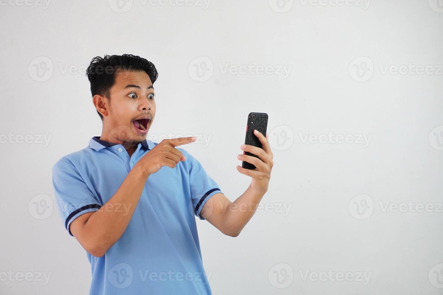 portrait asian man shocked holding phone and pointing at the phone with a finger wearing blue polo t shirt isolated on white background photo