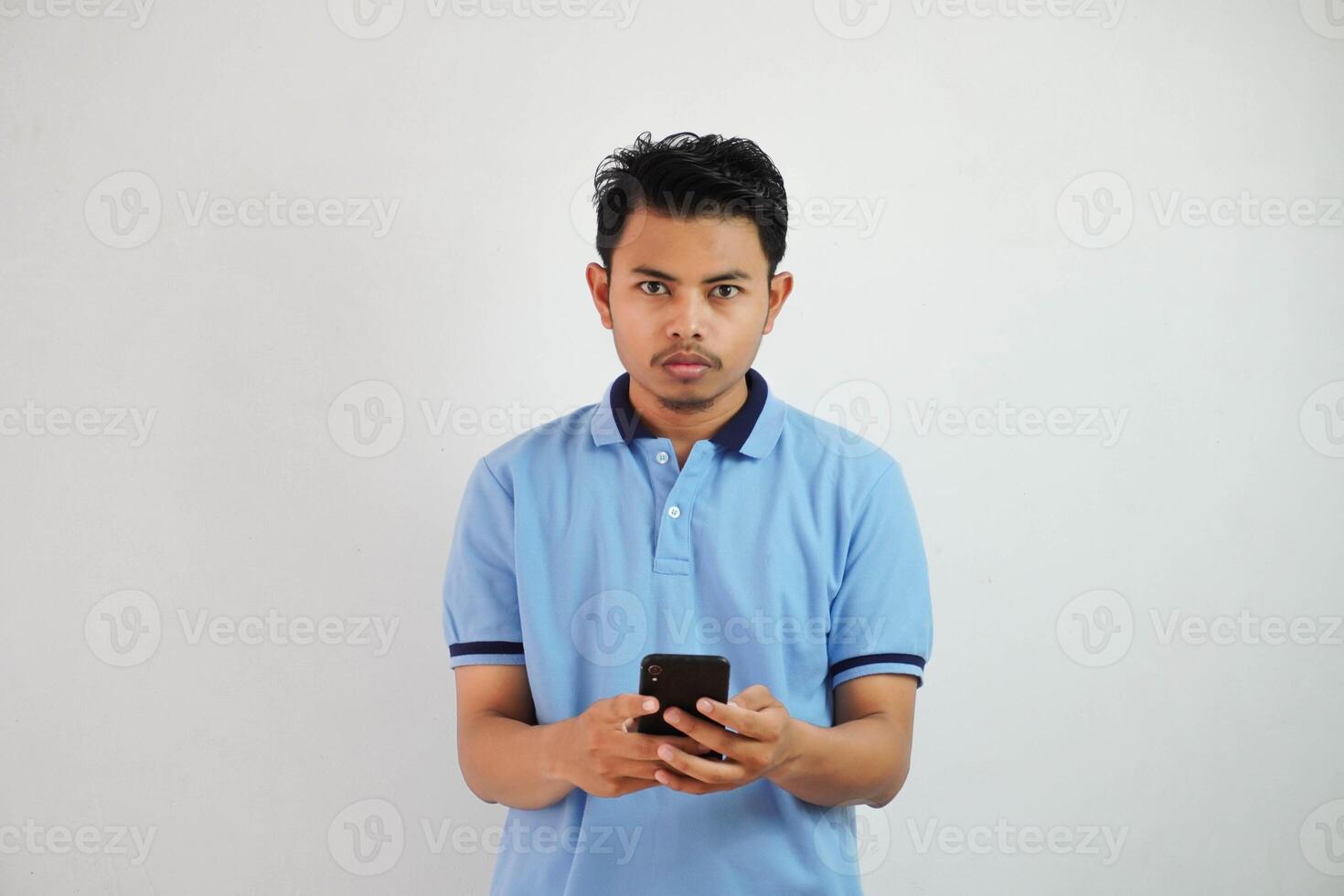 joven asiático hombre en pie participación un teléfono con un enojado y desaprobando expresión vistiendo azul t camisa aislado en blanco antecedentes foto