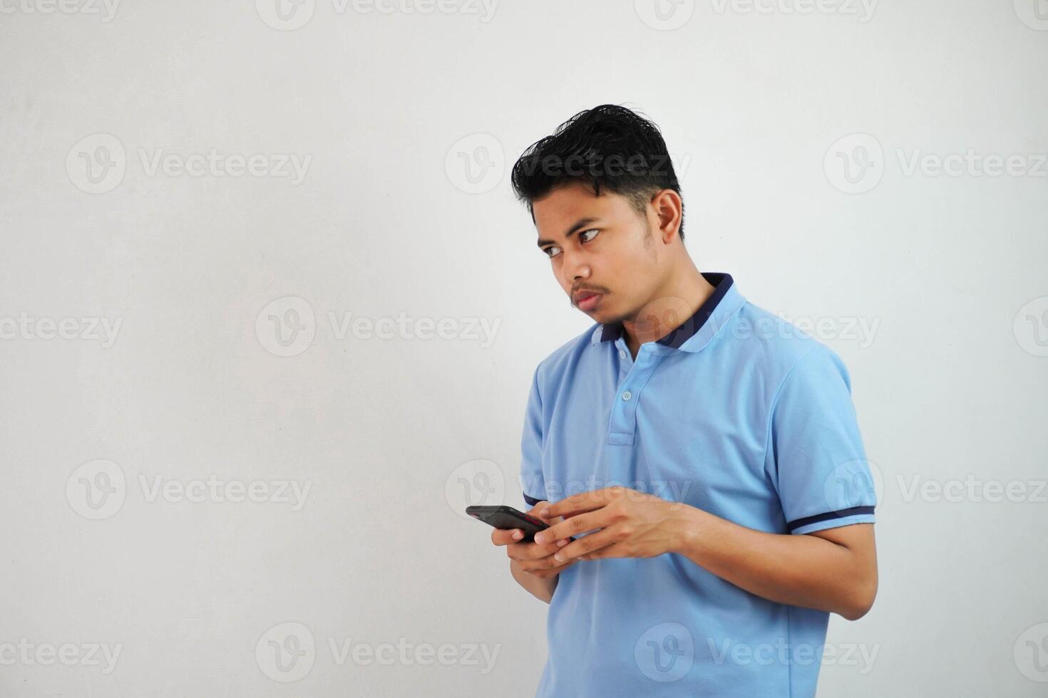 joven asiático hombre en pie participación un teléfono con un enojado y desaprobando expresión vistiendo azul t camisa aislado en blanco antecedentes foto