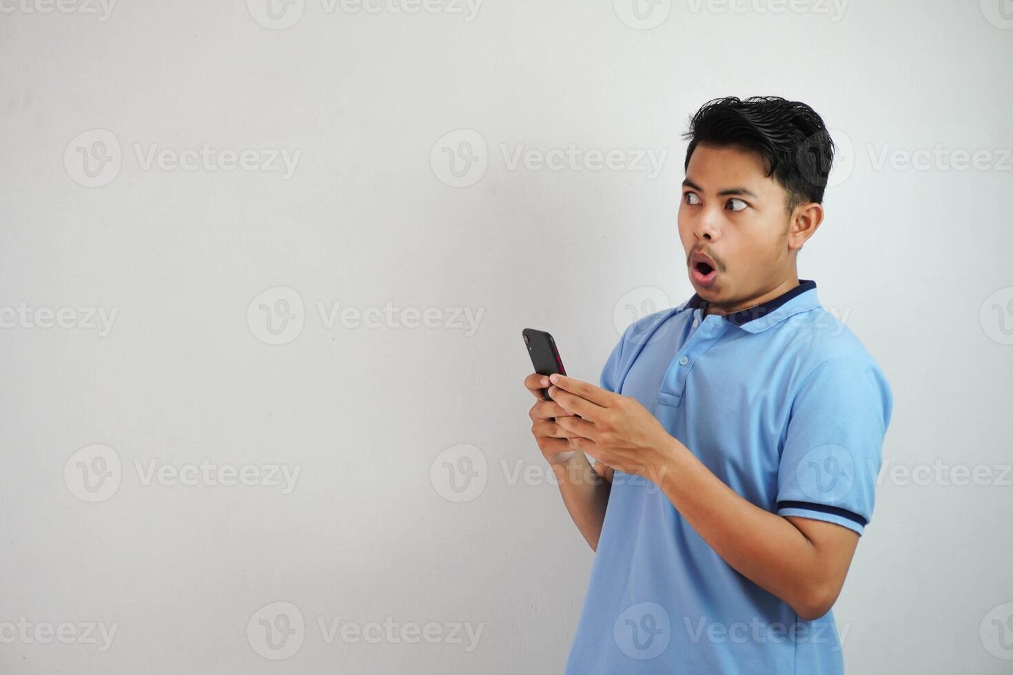 shocked young asian man holding phone and open mouth wearing blue t shirt isolated on white background photo