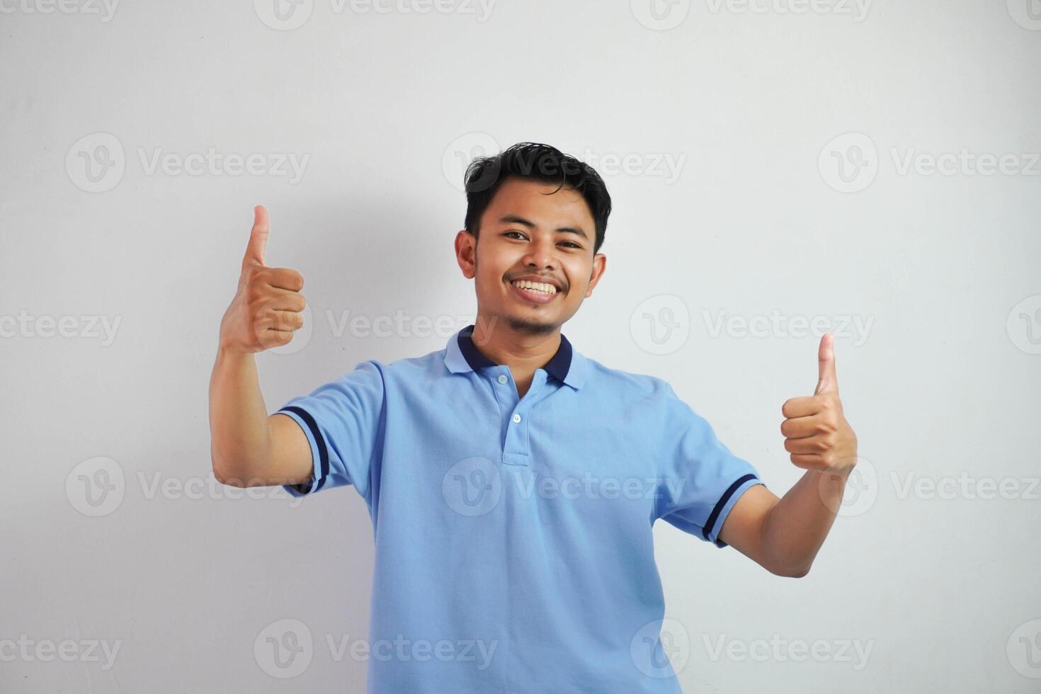Portrait of cheerful asian man in wearing blue t shirt smiling and showing thumbs up at camera isolated over white background photo