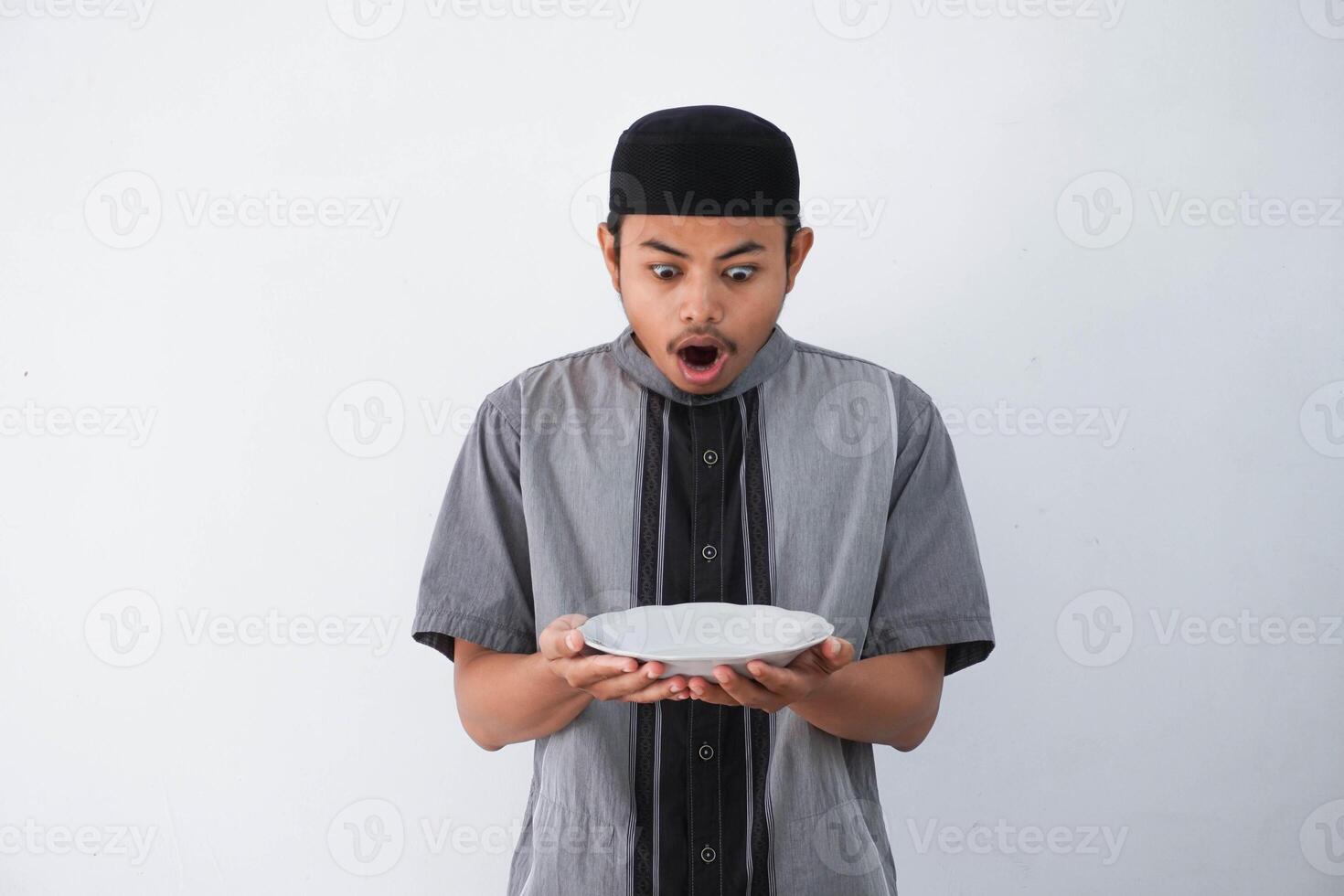 shocked young Asian muslim man showing excited expression while holding empty dinner plate wearing grey muslim clothes isolated on white background photo