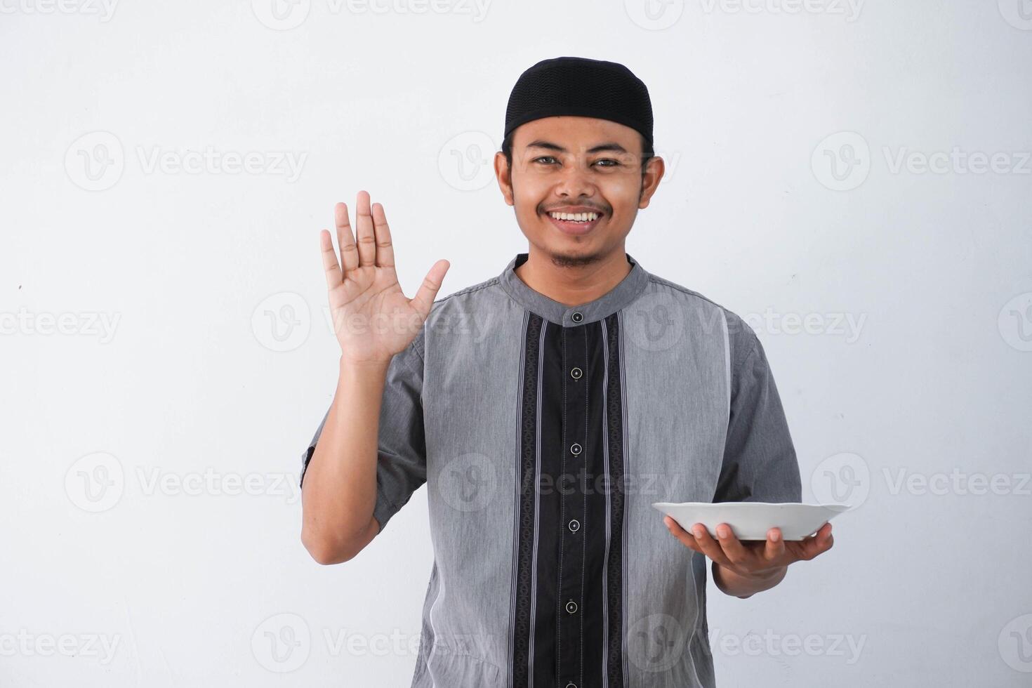 smiling or happy young Asian muslim man showing excited expression while holding empty dinner plate wearing grey muslim clothes isolated on white background photo