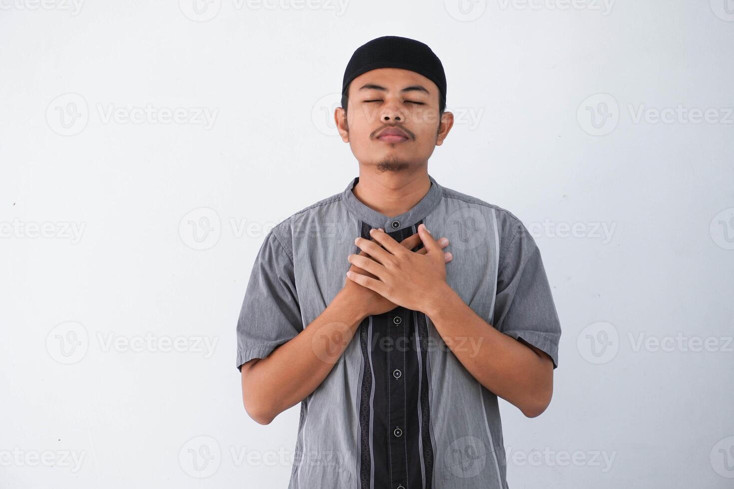 young Asian muslim man wearing grey muslim clothes with cap hands on chest with closed eyes and grateful gesture on face isolated on white background. health concept. photo