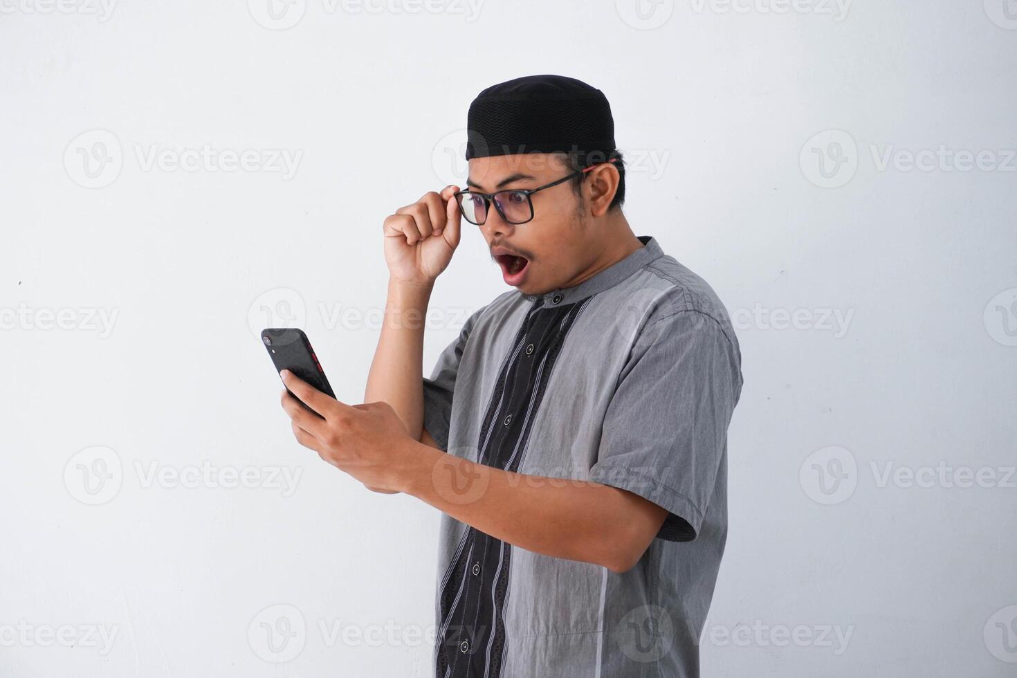 shocked asian muslim man holding phone and glasses wearing grey muslim clothes isolated on white background photo