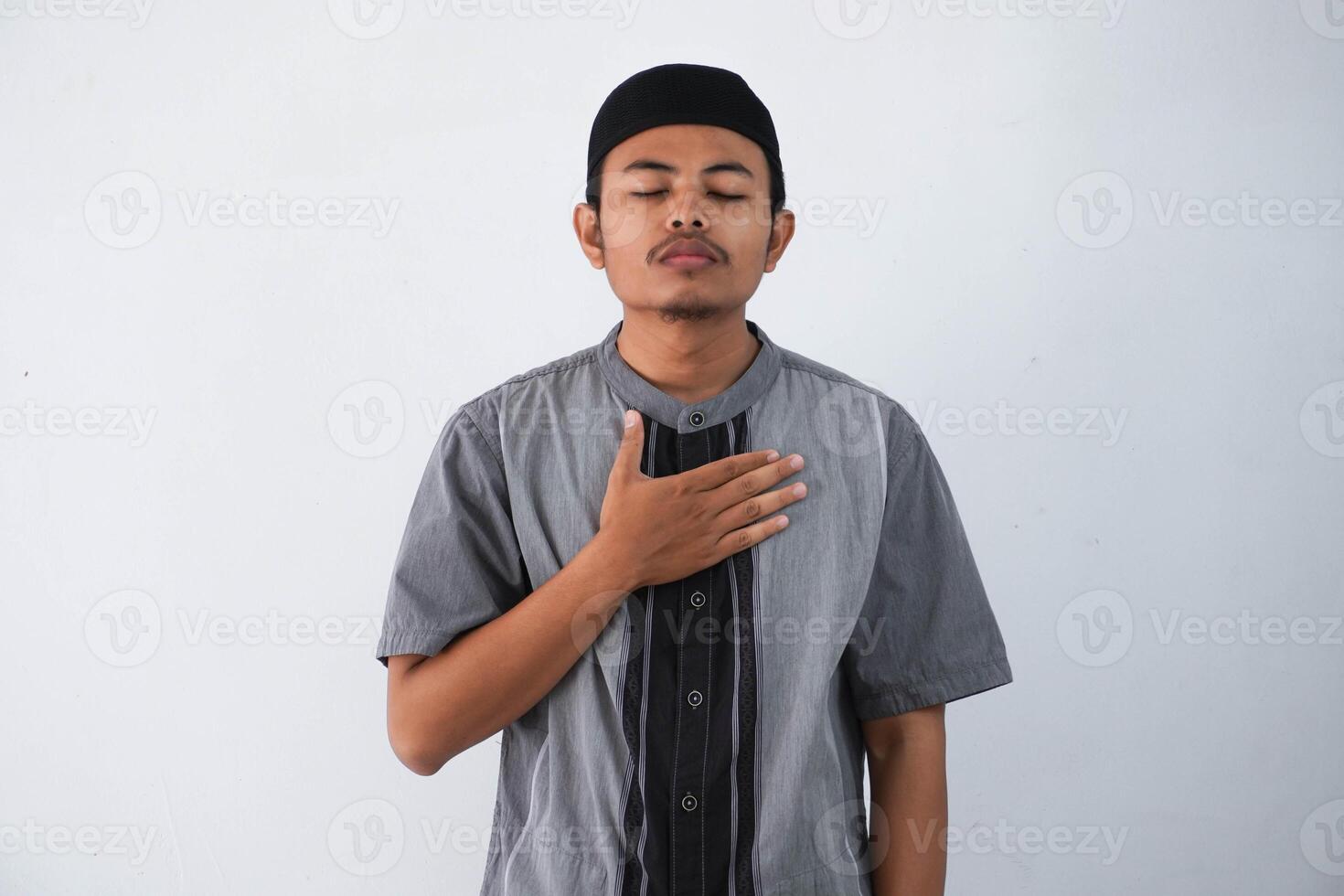 young Asian muslim man wearing grey muslim clothes with cap hands on chest with closed eyes and grateful gesture on face isolated on white background. health concept. photo