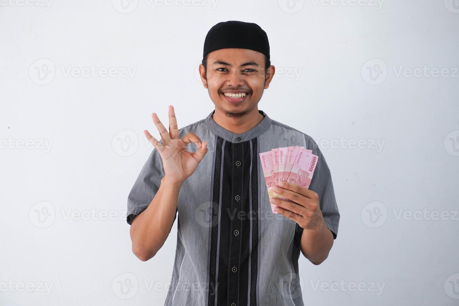 sonriente o contento joven asiático musulmán hombre dedo bueno gesto participación papel dinero regalo thr desde familia Ramadán eid al-fitr hora vistiendo gris musulmán ropa aislado en blanco antecedentes foto