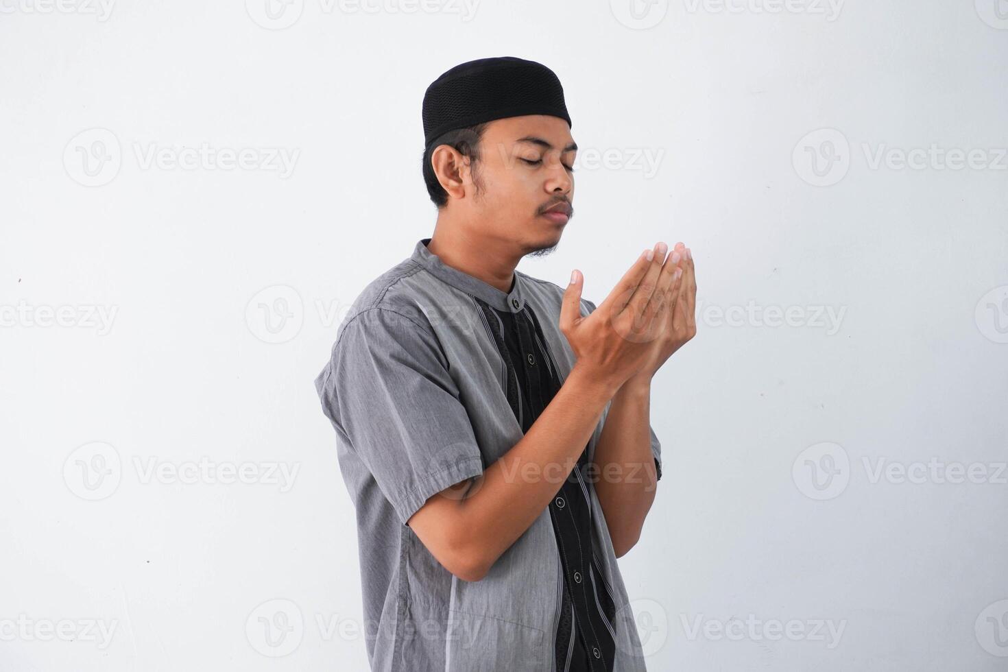 Religious young asian muslim man with close eyes praying, holding palms face up, whispering pray, isolated on white background. Religion islam, believing concept photo