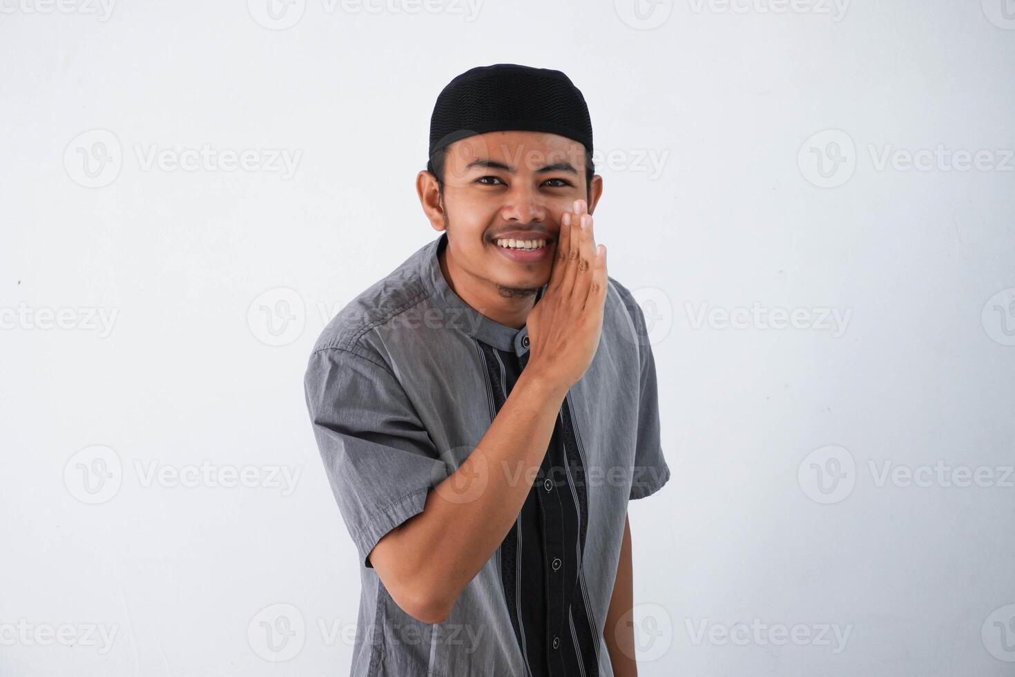 young Asian Muslim man wearing grey muslim cloth, hand beside mouth whispering secrets or gossips isolated on white background photo