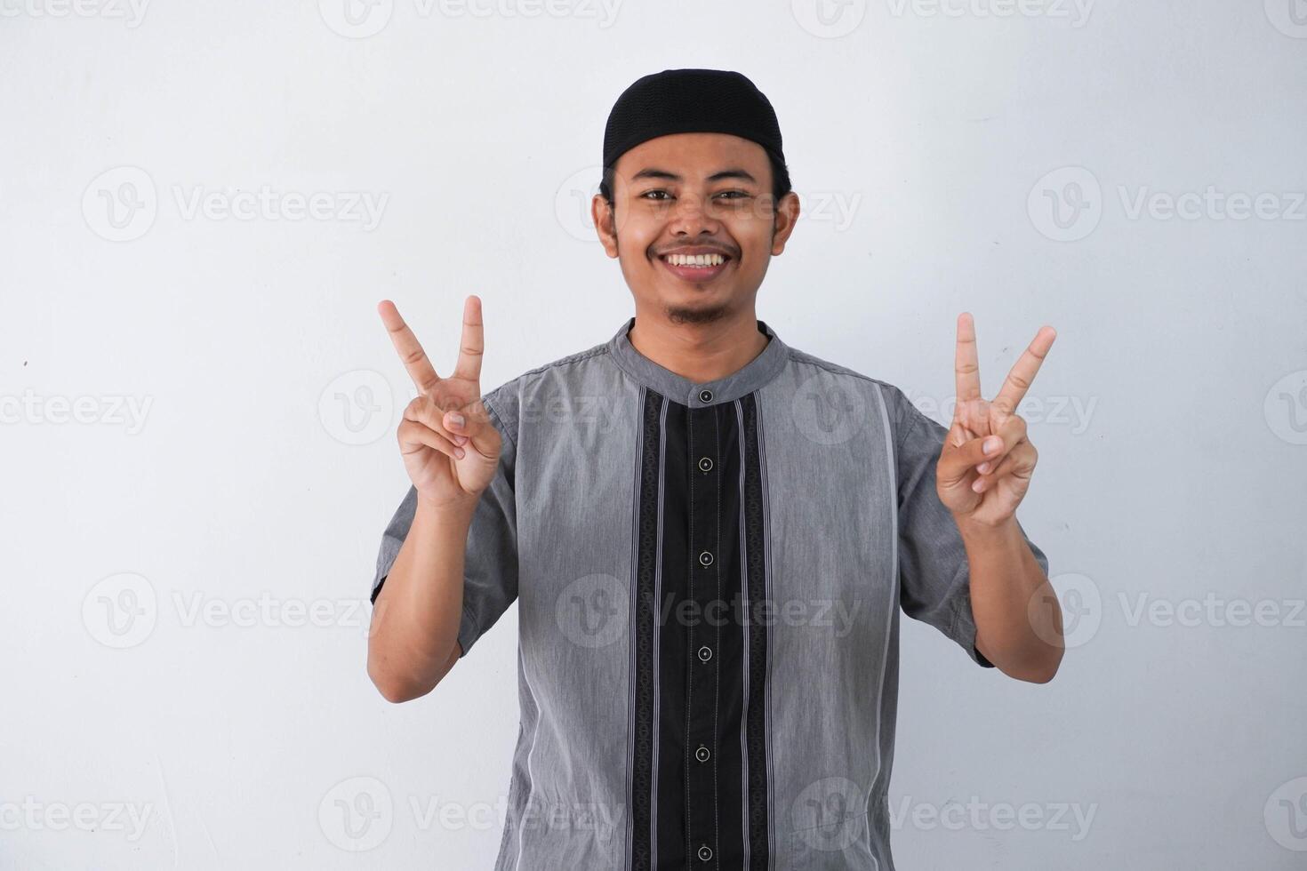 smiling or happy young asian muslim man showing pointing up with fingers number two while confident and showing peace sign with fingers wearing koko clothes isolated on white background photo