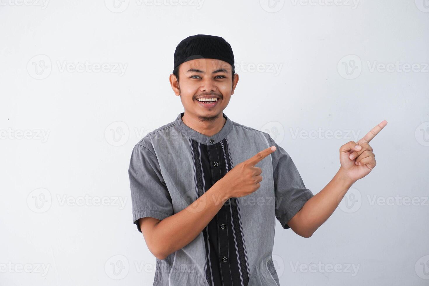 happy young asian muslim man pointing something on his side with his both hand wearing grey muslim clothes isolated on white background photo