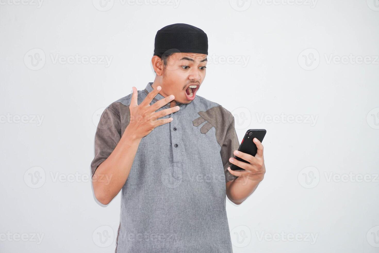 angry or shocked young asian muslim man looking a phone with open arms and open mouth wearing muslim clothes isolated on white background photo