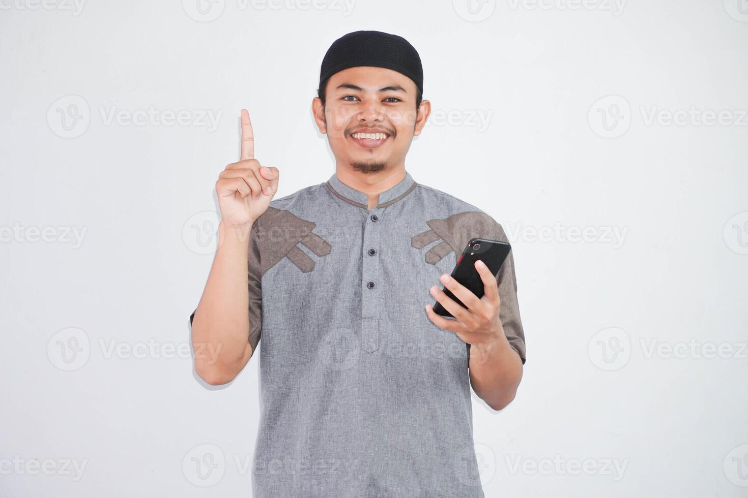 sonriente joven asiático musulmán hombre participación móvil teléfono y señalando dedo arriba, creando genio soluciones vistiendo gris musulmán ropa aislado en blanco antecedentes foto
