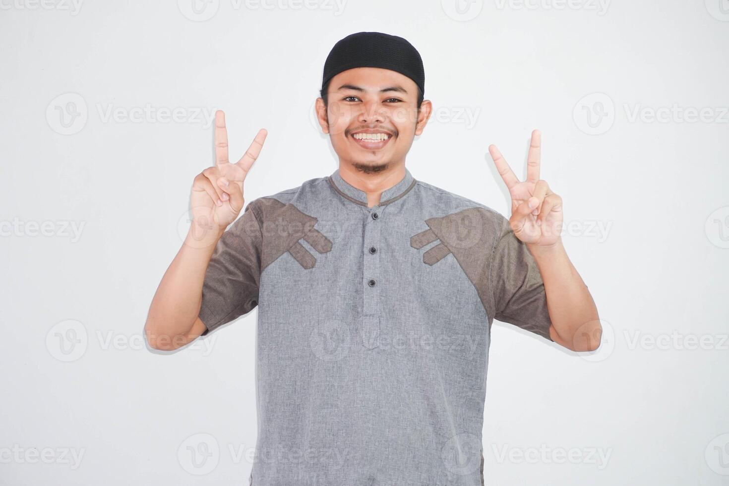 smiling or happy young asian muslim man showing pointing up with fingers number two while confident and showing peace sign with fingers wearing koko clothes isolated on white background photo