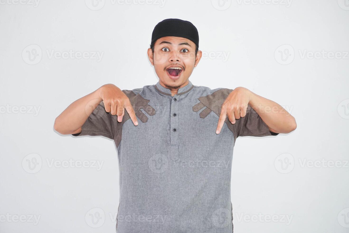 shocked young asian muslim man open mouth and amazed, looking and pointing downwards in disbelief and surprise wearing grey koko clothes isolated on white background photo