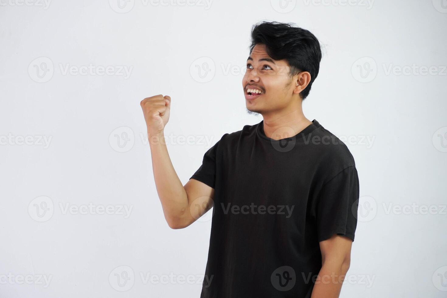 Confident cheerful handsome asian man lifting hands up wearing black t shirt winner gesture clenching fists. feels happiness show fist up success isolated on white background photo