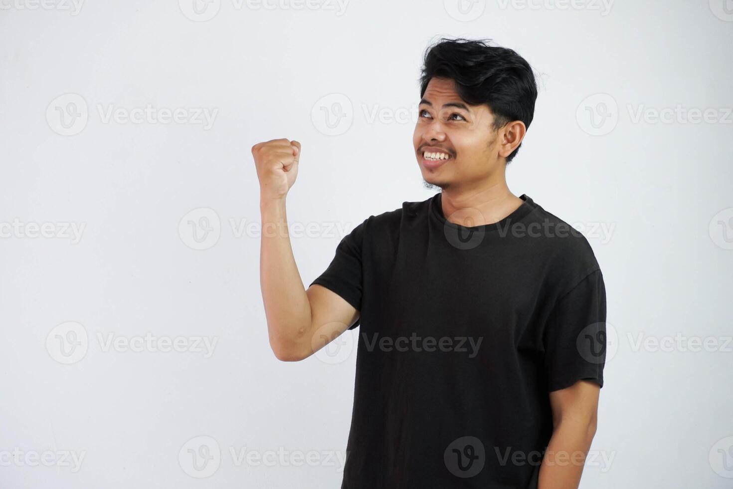 Confident cheerful handsome asian man lifting hands up wearing black t shirt winner gesture clenching fists. feels happiness show fist up success isolated on white background photo