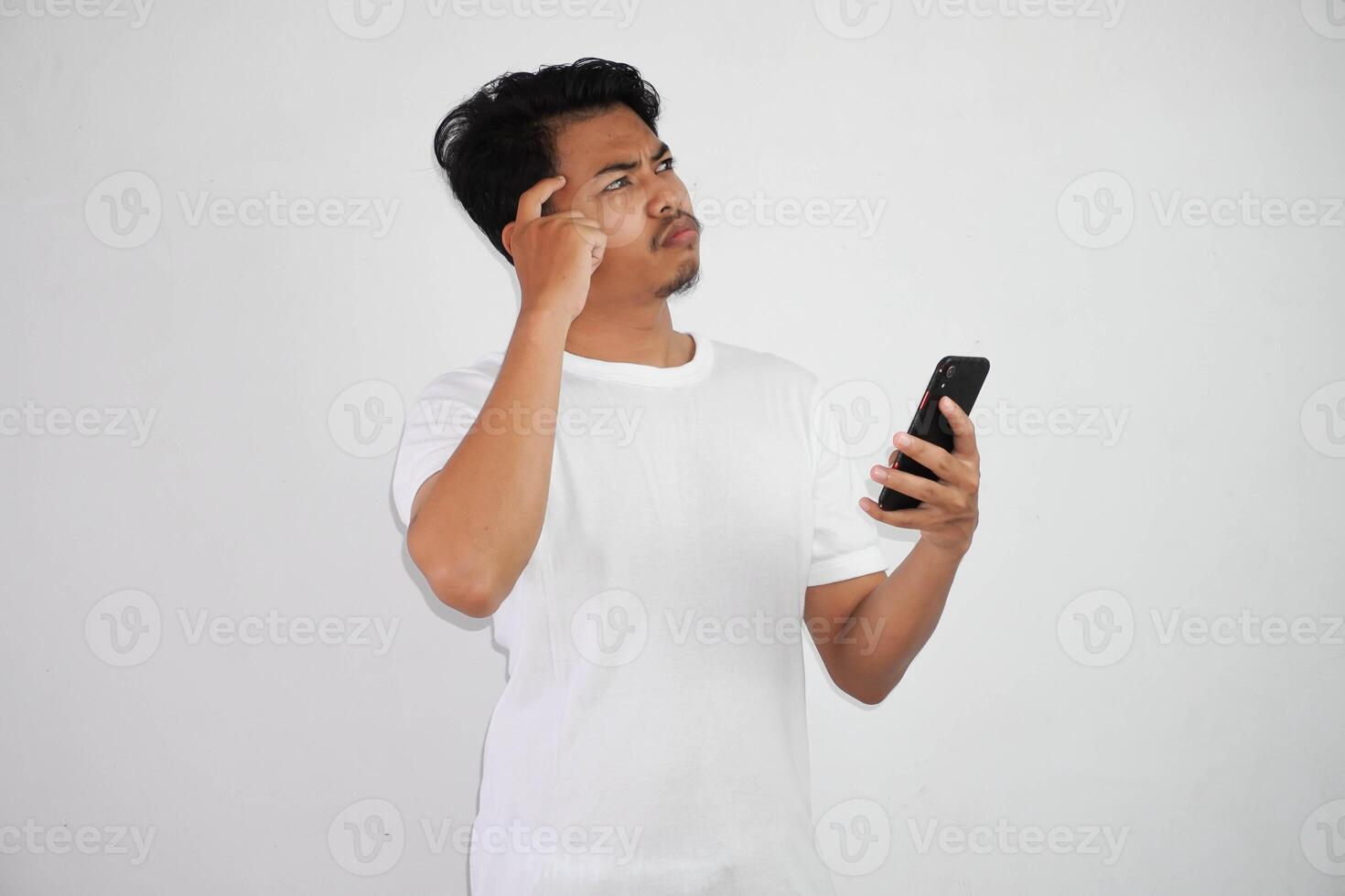 thinking young asian man holding mobile phone pointing head thinking gesture, looking for idea wearing white t shirt isolated on white background photo
