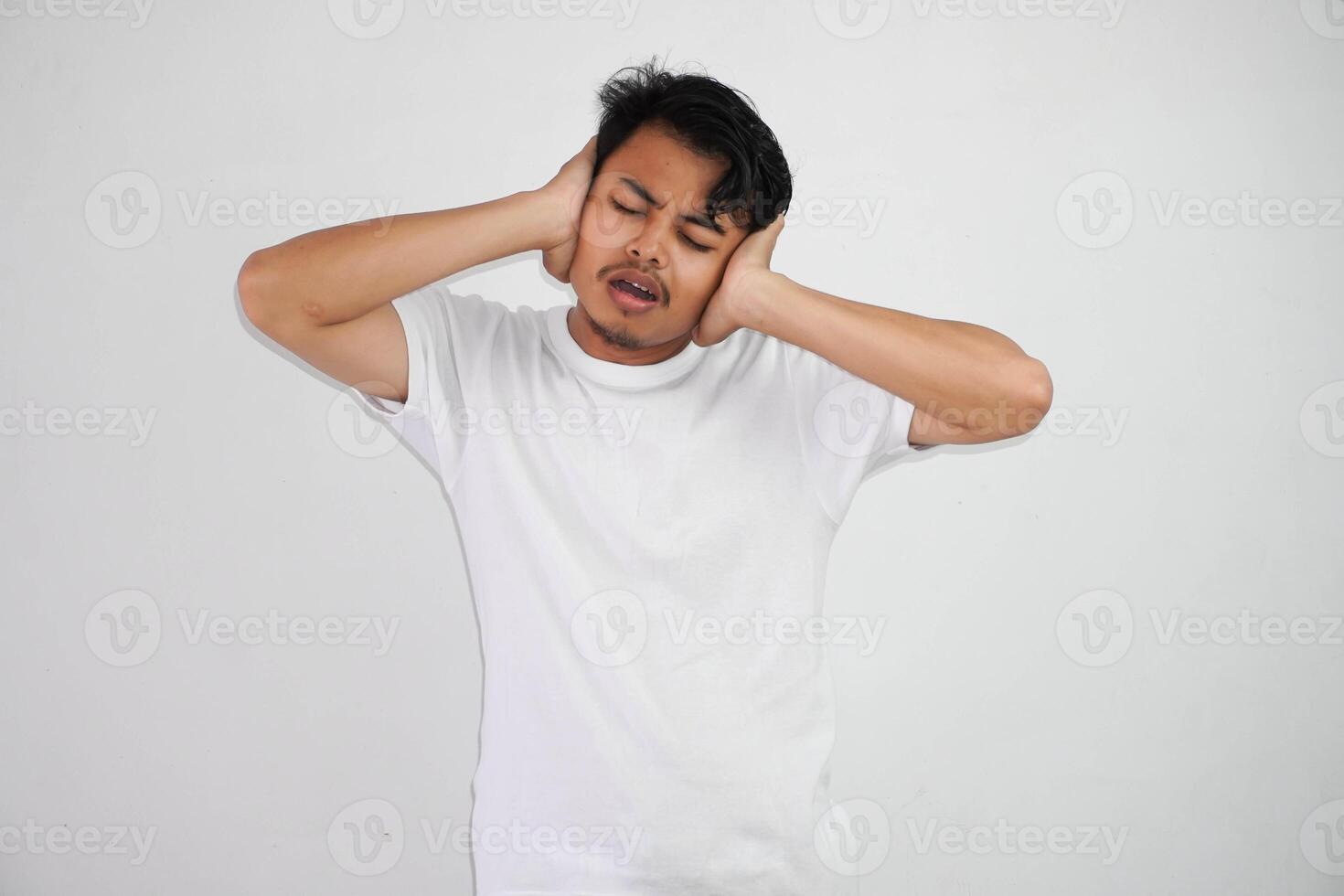 Frustrated young Asian man covering ears with hands and keeping eyes closed wearing white t shirt isolated on white background photo