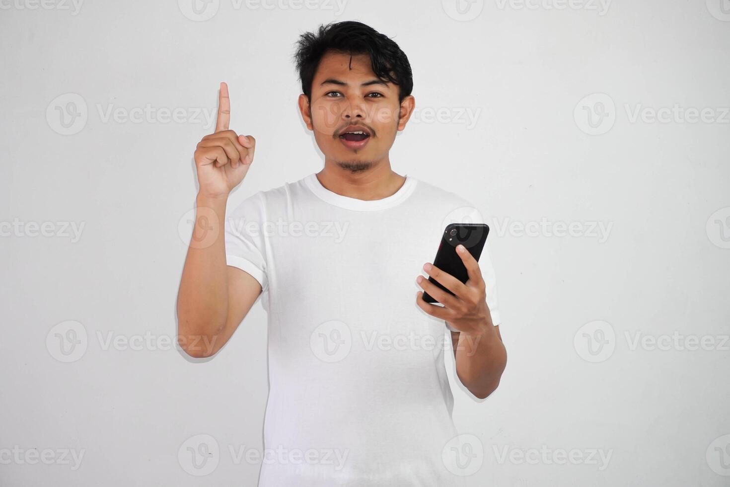 Excited young Asian man pointing fingers up having a good idea with holding mobile phone wearing white t shirt isolated on white background photo