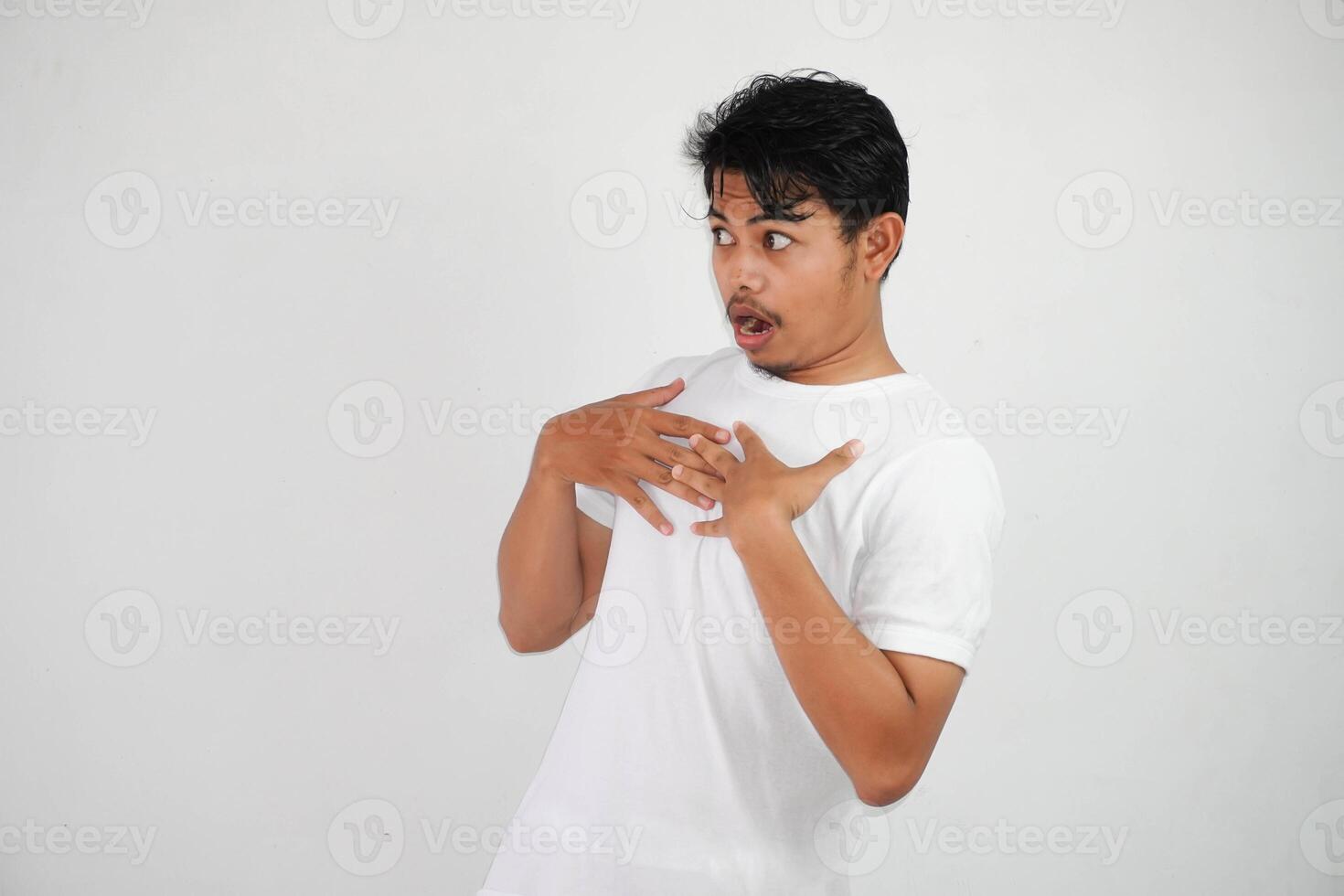 conmocionado joven asiático hombre con abierto boca y poner un mano en pecho, mirando lejos vistiendo blanco t camisa aislado en blanco antecedentes foto