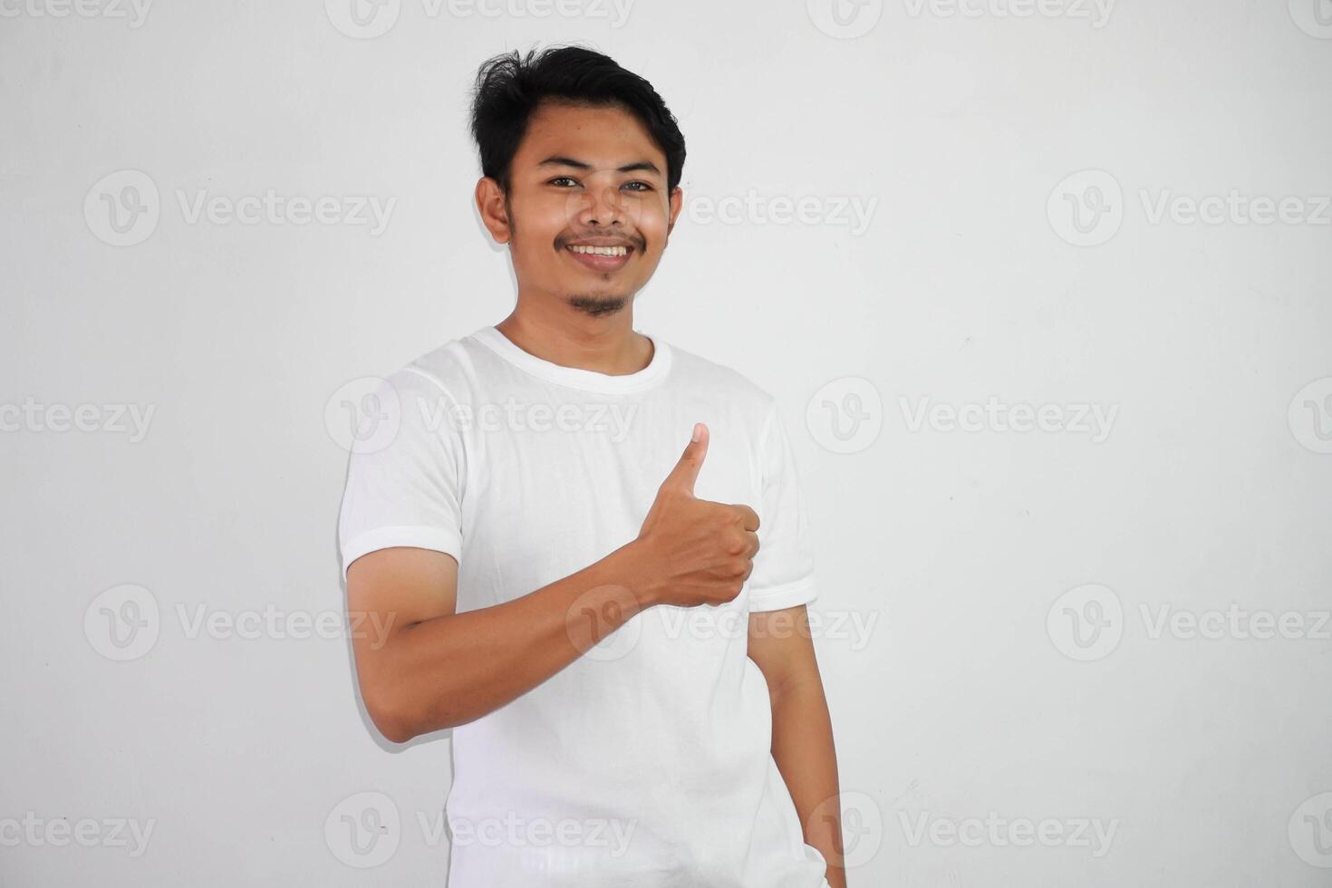 retrato de alegre asiático hombre en vistiendo blanco t camisa sonriente y demostración pulgares arriba a cámara aislado terminado blanco antecedentes foto