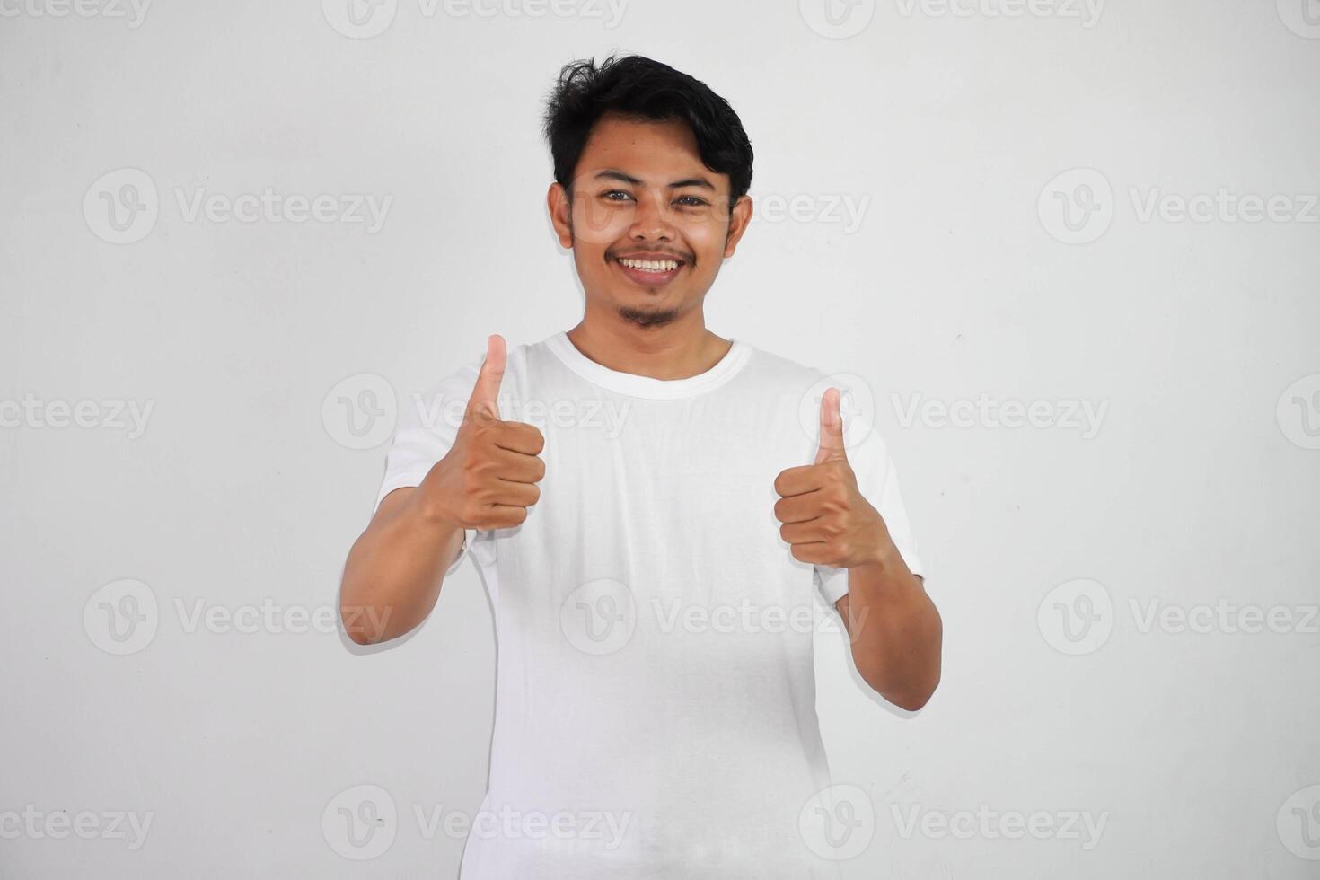 retrato de alegre asiático hombre en vistiendo blanco t camisa sonriente y demostración pulgares arriba a cámara aislado terminado blanco antecedentes foto