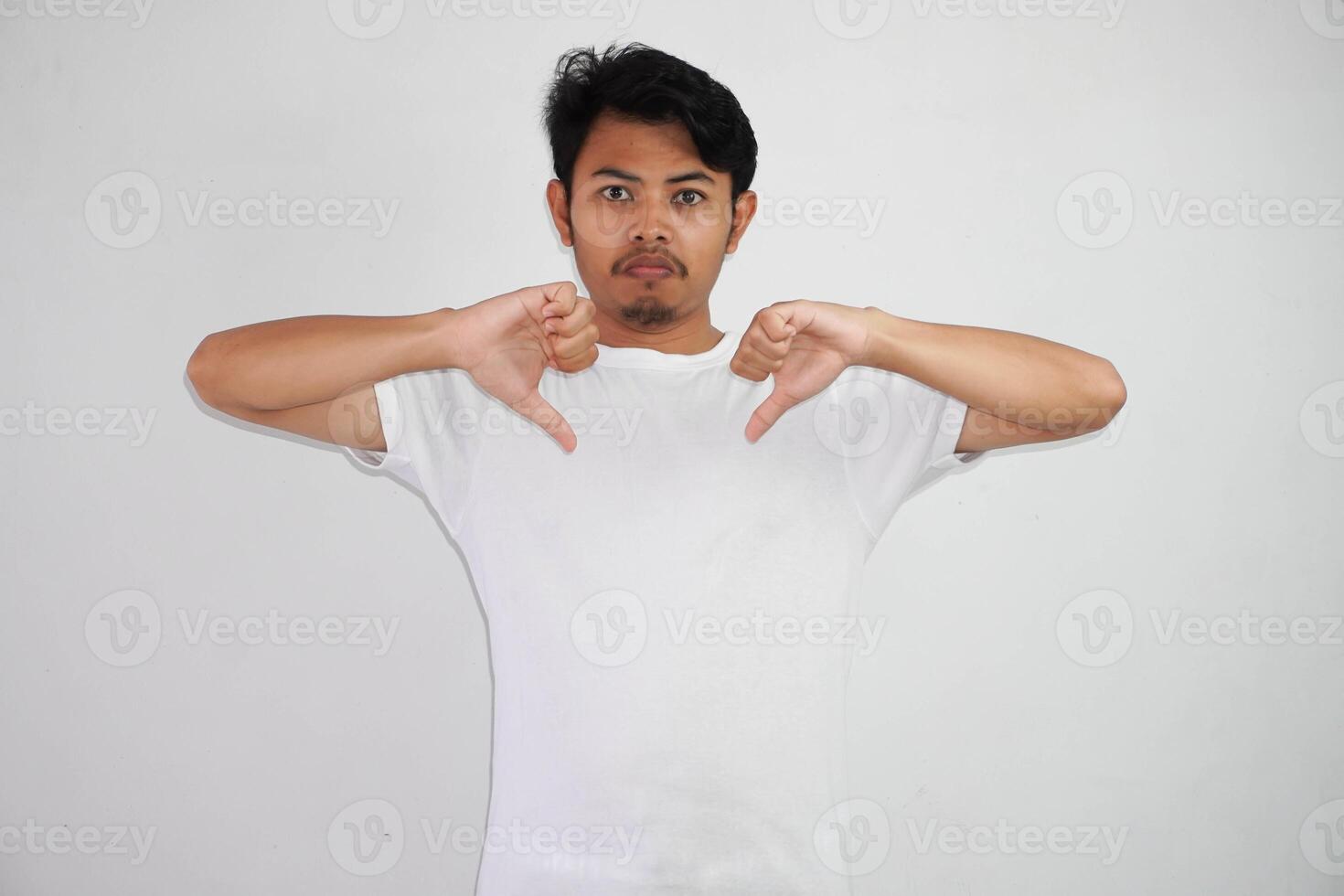 enojado joven asiático hombre demostración rechazo y negativo con pulgares abajo gesto vistiendo blanco t camisa aislado en blanco antecedentes foto