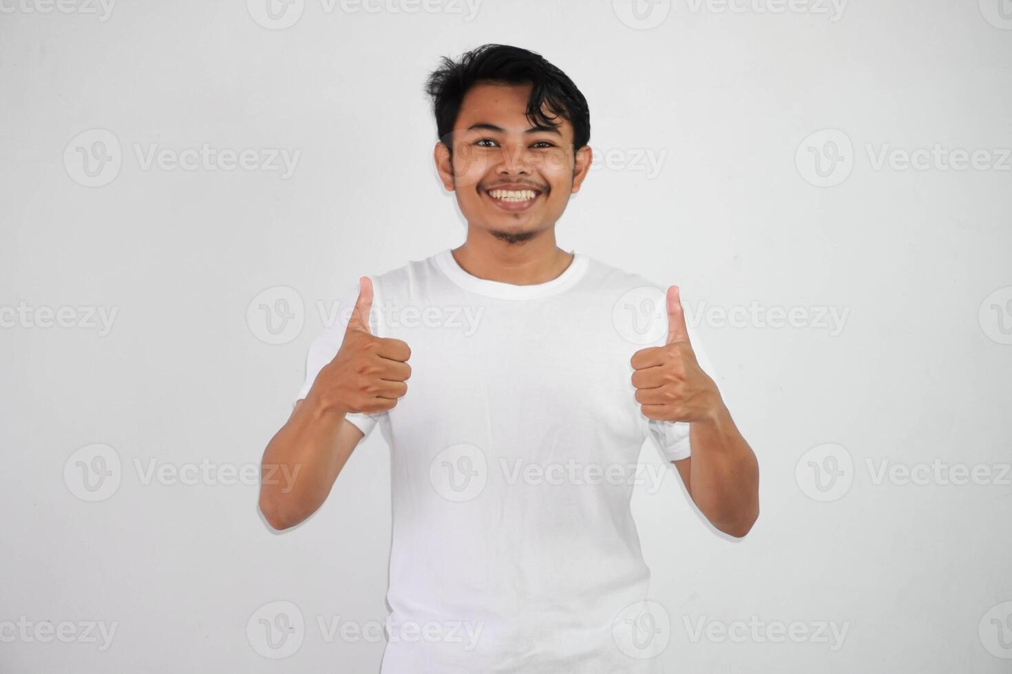 retrato de alegre asiático hombre en vistiendo blanco t camisa sonriente y demostración pulgares arriba a cámara aislado terminado blanco antecedentes foto