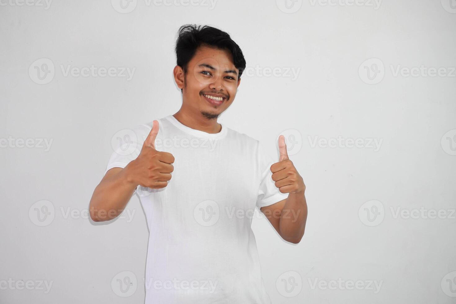 retrato de alegre asiático hombre en vistiendo blanco t camisa sonriente y demostración pulgares arriba a cámara aislado terminado blanco antecedentes foto