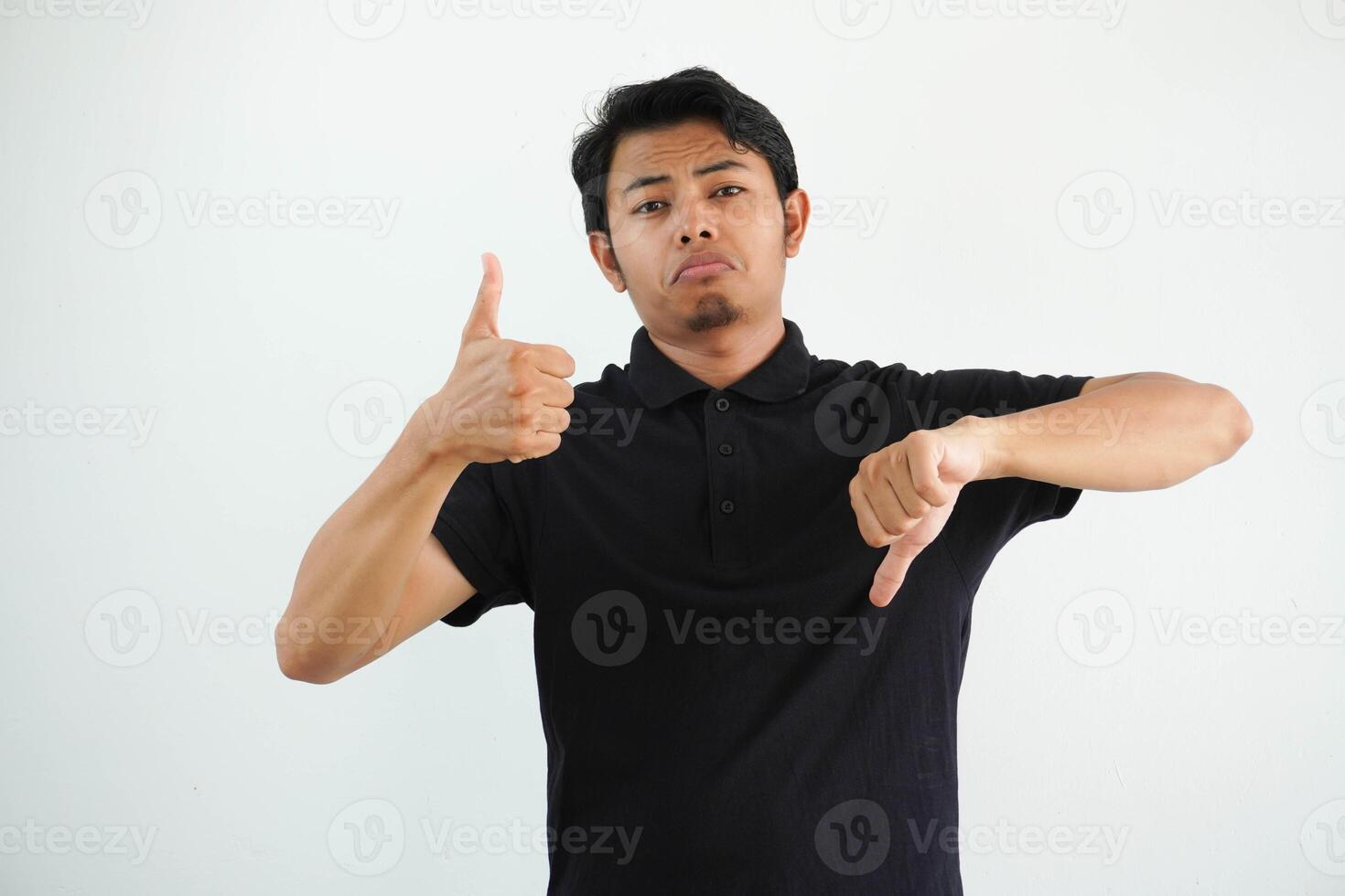 joven asiático hombre en negro polo t camisa, blanco estudio fondo demostración pulgares arriba y pulgares abajo, difícil escoger concepto foto