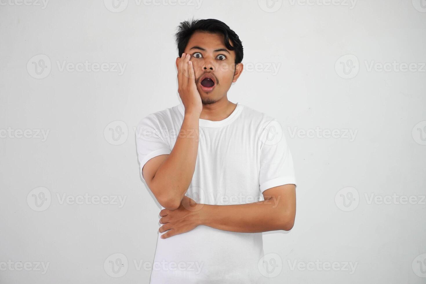conmocionado joven asiático hombre en pie mano en mejilla con abierto boca vistiendo blanco t camisa aislado en blanco antecedentes foto