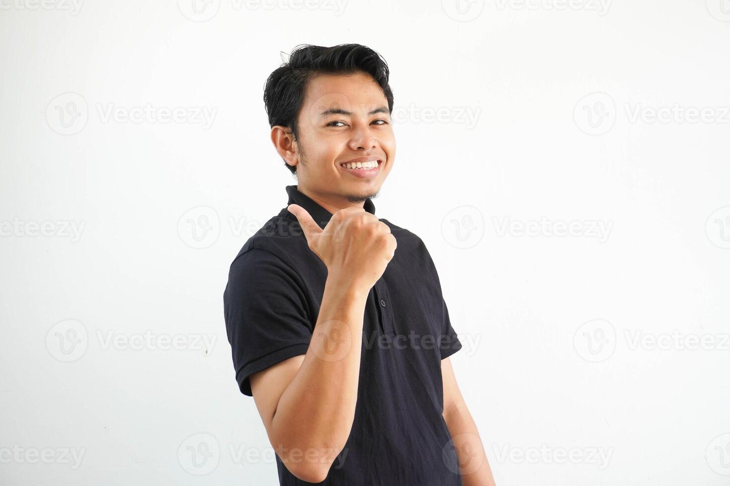 smiling young asian man against a vibrant white studio background, thumb pointing to the right side, with thumb finger away, laughing and carefree, wearing black polo t shirt. photo