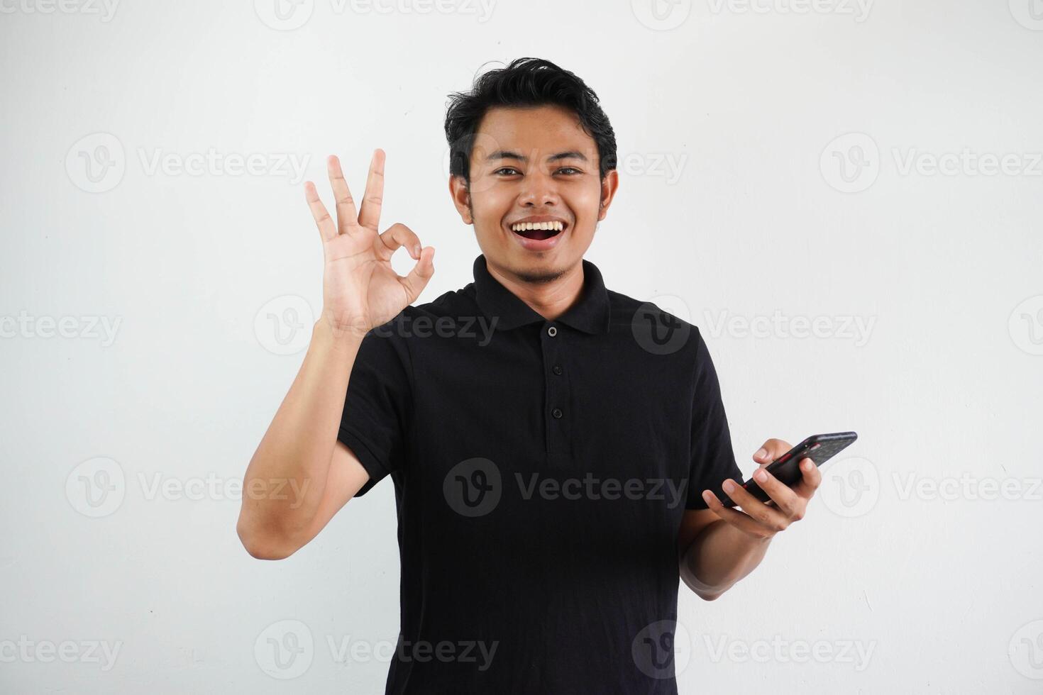 joven asiático hombre sonriente confidente y dar Okay dedo firmar mientras participación un móvil teléfono vistiendo negro polo t camisa aislado en blanco antecedentes foto