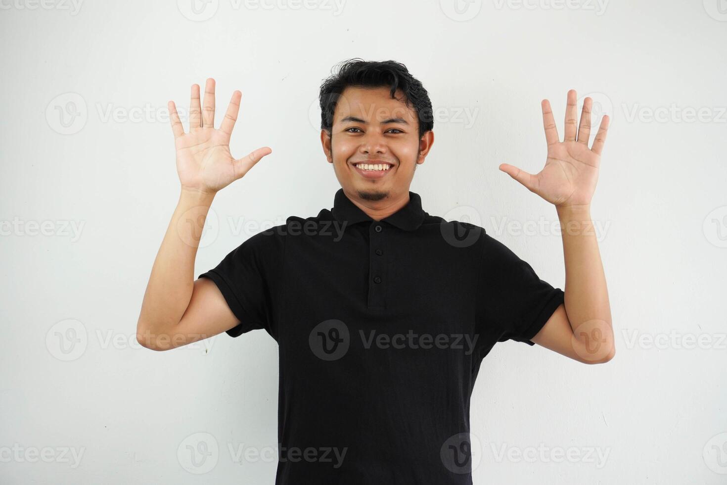 sonriente o contento joven asiático hombre con elevado ambos manos arriba vistiendo negro polo t camisa aislado en blanco antecedentes foto