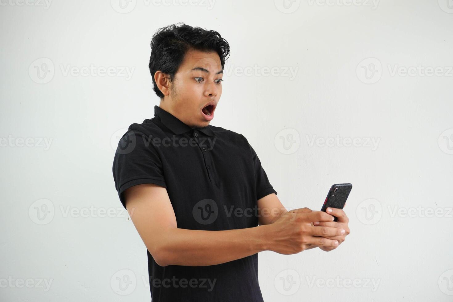 Shocked young asian man wearing polo t shirt looking at his phone with surprise expression, with mobile device learning unbelievable news, amazed at promo sale isolated on white background photo